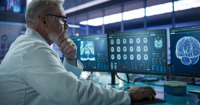 Senior Male Neuroscientist Using Desktop Computer To Analyze MRI Scans Of Brain In Medical Research Center Laboratory. Caucasian Surgeon Studying CT Scan Of Patient Suffering From Brain Tumor.