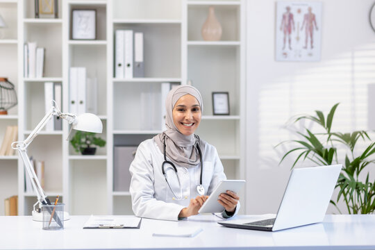 Portrait of Muslim female doctor in hijab, female doctor with tablet computer looking at camera with smile, working inside medical office, wearing white medical coat, sitting at table with laptop.