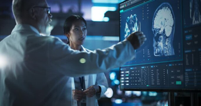 Neuroscience Laboratory Meeting: Female Asian Scientist And Male Caucasian Surgeon Discussing MRI Brain Scan On TV Display. Specialists Working On Medication For Rehabilitation After Brain Damage.