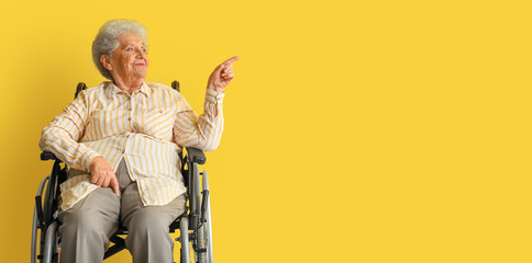 Senior woman in wheelchair pointing at something on yellow background with space for text
