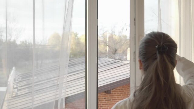 The woman parted the transparent tulle curtains and turned the handle on the PVC plastic window frame and opened the window wide to ventilate the room.