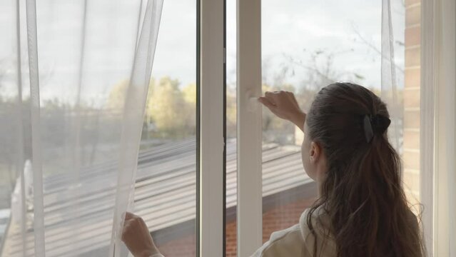 The woman parted the transparent tulle curtains, opened the window by turning the handle of the PVC plastic window frame, and stood at the window, looking out.