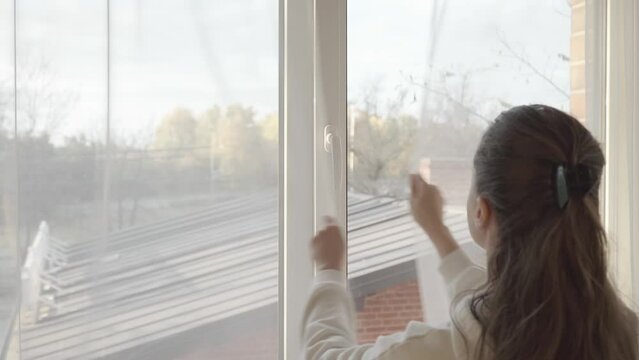 The woman tightly closes the wide-open window, turns the handle on the PVC plastic window frame and pulls the transparent tulle curtains.