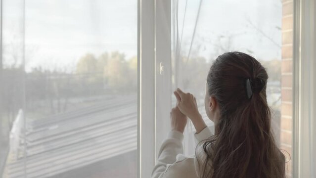The woman tightly closes the slightly ajar window, turns the handle on the PVC plastic window frame and pulls the transparent tulle curtains.