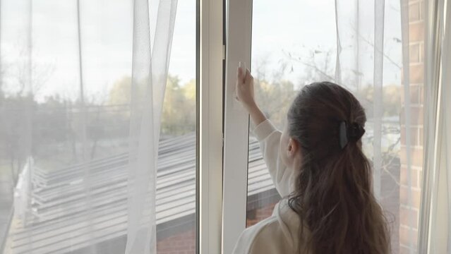 The woman parted the transparent tulle curtains and turned the handle on the PVC plastic window frame to slightly open the window and ventilate the room through a small crack.