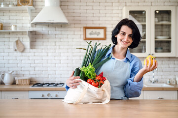 Smiling woman at home getting groceries out of shopping bag - obrazy, fototapety, plakaty