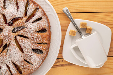 Sweet homemade plum cake with white ceramic dish, cup, saucer and knife and fork on wooden table, macro, top view.