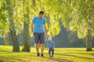 Young adult father and baby boy walking on green grass through tree alley at park. Spending time...