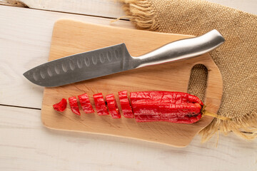 One red hot pepper cut into pieces on a wooden kitchen board with a metal knife, macro, top view.