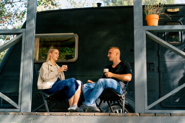 Fototapeta na wymiar A young handsome guy and a girl are sitting on the veranda of a motorhome in camper drinking tea and talking enthusiastically