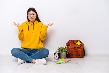 Young Chinese student woman sitting on the floor isolated on white wall having doubts while raising hands