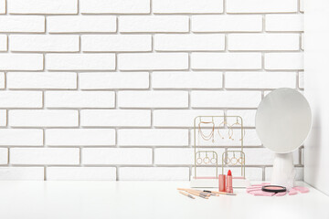 Different makeup products and mirror on dressing table near light brick wall in room