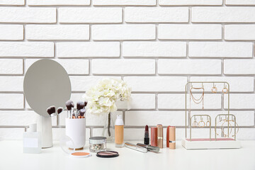 Different makeup products, mirror and beautiful flowers on dressing table near light brick wall in...