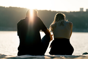 Silhouette of couple in love sitting on the sandy bank of the river admiring the setting sun and...