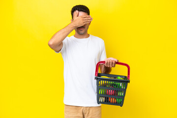 Young handsome man holding a shopping basket full of food over isolated yellow background covering...