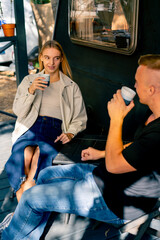 A young beautiful girl drinks tea and talks with her boyfriend while sitting on the terrace near a motorhome in camper