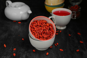 Bowl with red dried goji berries and cup of hot tea on black background