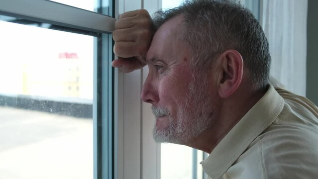 Sad tired lonely disappointed older senior man at home alone. Unhappy grandfather looking sadly outside window experiencing grief relative death bad news. Stressed elder man suffering from loneliness
