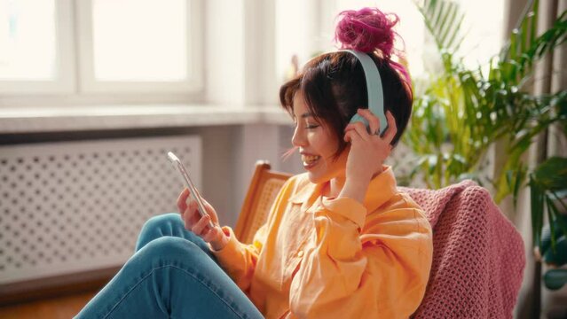 Portrait of a young bright Asian woman with pink strands of hair listening to music or online training in headphones sitting in an armchair and relaxing in the living room.