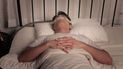 Asian man using CPAP mask and machine for sleep apnea treatment at home
