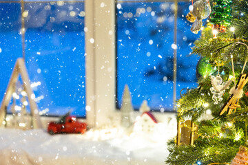 Fototapeta premium Cozy Christmas decor with houses, a truck on the windowsill with snowfall near the Christmas tree.