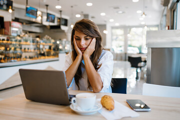 Beautiful and happy young woman sitting and eating delicious rolls in bakery or fast food while she using laptop computer. She is serious and tired. - Powered by Adobe