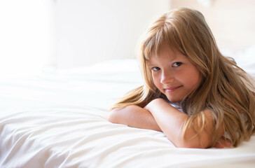 Fototapeta premium A little happy girl with long hair is lies on a white comfortable bed look front to the camera at the morning. Copy space.It's perfect for projects related to family, childhood, or relaxation.