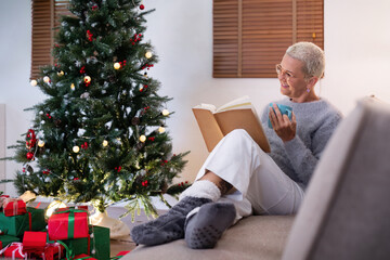 Obraz na płótnie Canvas Smiling elderly woman with a cup of tea and a book relax in side of a decorated Christmas tree in living room