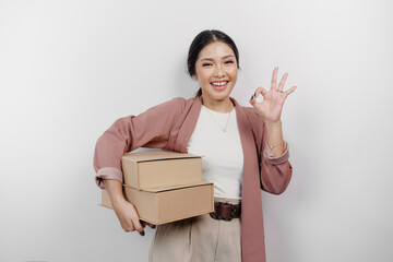 Excited Asian woman employee wearing a cardigan giving an OK hand gesture while holding stack of cardboard boxes, isolated by a white background