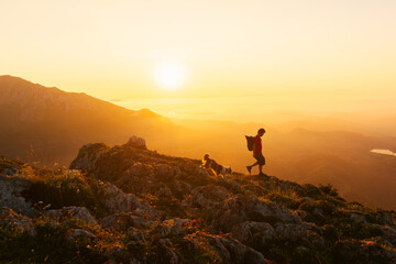 young man in silhouette descending a mountain peak at sunset with his dog. Sport, adventure and hiking in the mountains.