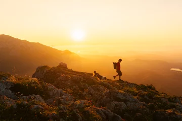 Poster young man in silhouette descending a mountain peak at sunset with his dog. Sport, adventure and hiking in the mountains. © Alberto