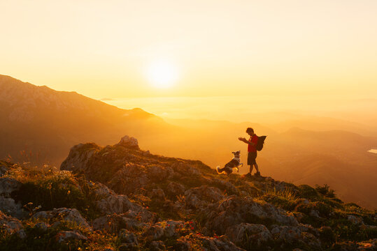 Mountaineer man with backpack plays with his border collie dog while hiking in the mountains at sunset. Mountain sports and adventure