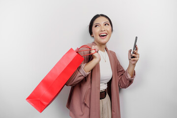 Naklejka premium A happy beautiful young Asian woman employee wearing cardigan is carrying shopping bag and her smartphone, isolated on white background