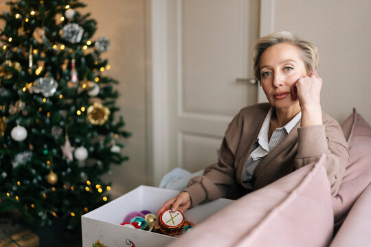Portrait of elegant middle-aged woman holding in hand festive xmas tree toy, sitting on couch. Preparation for holidays, decorates Christmas tree for New Year. Concept of home festive atmosphere.