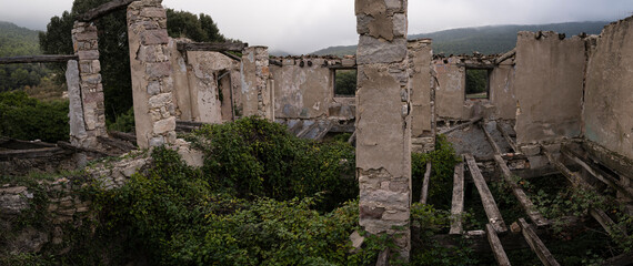 Abandoned Farmhouse in Ruins Amidst Overgrown Greenery with Forest and Mountains in the Background on a Cloudy Day