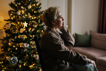 Side view of elegant woman talking on smartphone in dark room by illuminated Christmas tree. Happy female enjoying pleasant phone call, congratulating with New Year holidays, telling wishes.