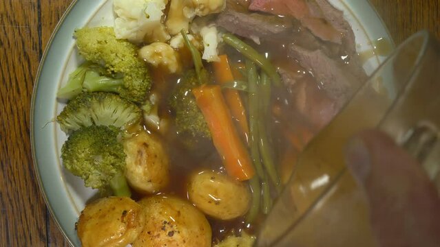 Closeup Overhead Shot Of Steaming Gravy Being Poured From A Measuring Jug Over A Dinner Plate Of Traditional Roast Beef, Vegetables And Yorkshire Pudding.