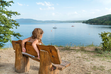 Kid sitting on a bench high above a blue lake with stunning view . 