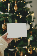 Hand holding empty greeting card on background of stylish decorated christmas tree with golden...