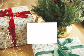 Empty greeting card on stylish wrapped christmas gift and fir branches with festive christmas lights on wooden table. Christmas card mock up. Space for text. Season greetings postcard template © sonyachny