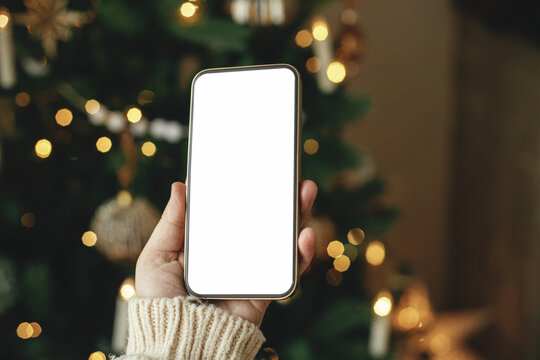 Hand holding smartphone with empty screen against stylish festive christmas tree with golden lights.Christmas phone mock up. Space for text. Christmas advertising, smartphone app template