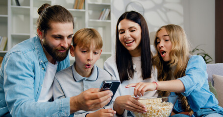 Happy young family relaxing on couch with adorable father daughter and son mother hand using smartphone application together in living room,family smartphone addiction.