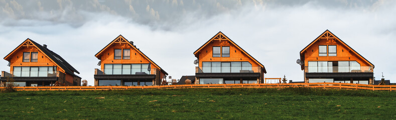 Fototapeta na wymiar Wide banner photo of the wooden houses in beautiful nature. Pretty landscape with modern wooden house surrounded by trees with colorful foliage and mountains. Runaway chalet cabins in the forest.