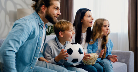 Family sitting on couch at home watching live broadcasting of soccer game together celebrating a...