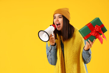 Young woman in winter clothes with Christmas gift shouting into megaphone on yellow background - obrazy, fototapety, plakaty