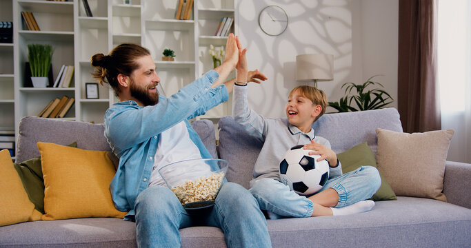 Funny blonde boy spending time together which father, watching soccer game on tv and emotionally reacting. Excited football fans celebrating victory of soccer team at home.