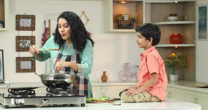 Indian mom cooking food and get food tested to her son sitting on the kitchen counter
