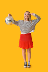 Beautiful young woman with disco ball showing victory gesture on yellow background