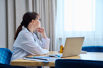 Confident serious mature woman doctor sitting at table with laptop computer