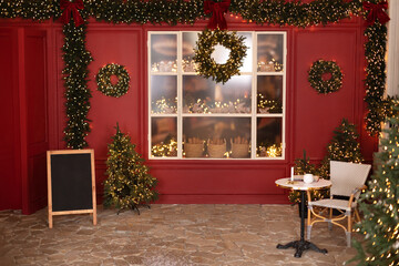 Christmas trees with lights garlands, store window. Red decorated coffee house for christmas. Xmas cafe terrace with table and chair. Cozy outdoor cafe on Xmas. New Year's decorations of the street.